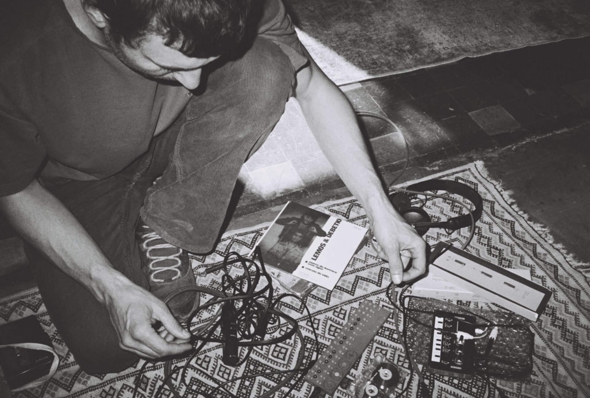 A black and white photo of man lying on the ground. He has wires in his hands. There are two rugs on the floor with headphones, cassette tape and music equipment on them.